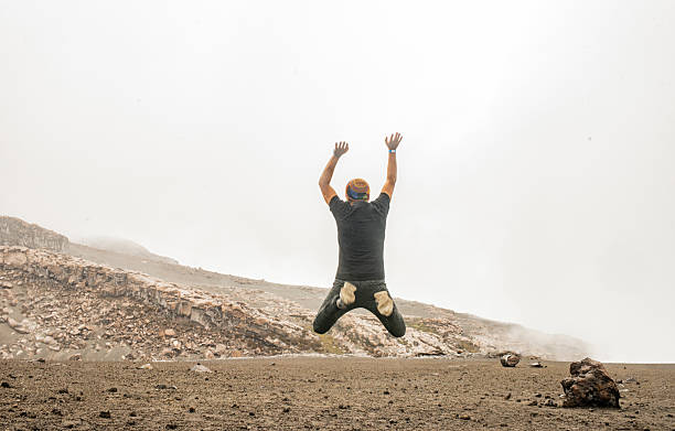 A person with their back to the camera jumps with their arms raised in an arid, mountainous volcanic environment surrounded by fog. Concept of freedom, energy, adventure, personal achievement, and connection with extreme nature.