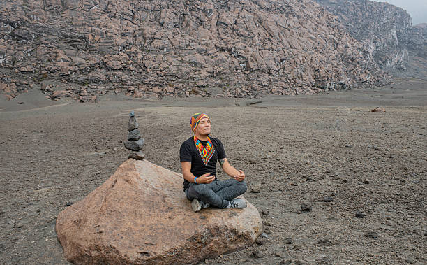 Young man sitting in a meditation position on a rock, with closed eyes and a calm expression, accompanied by a mound of stacked stones. Arid and mountainous setting, ideal for representing mindfulness, spirituality, and connection with nature.