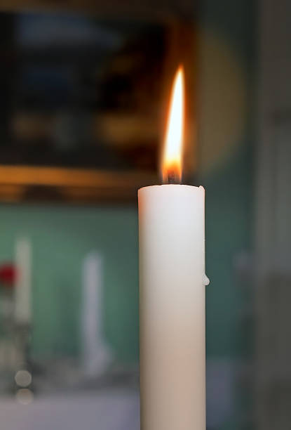 A lit candle with a bright, flickering flame stands against a subtly blurred indoor background. The warm light provides a sense of comfort, tranquility, and timeless beauty.