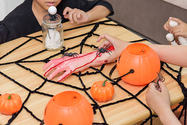 istock On a decorated table are pumpkin buckets, a jar, a spider web, a toy hand and toy spiders, children are visible creating festive crafts for halloween day 2233772679