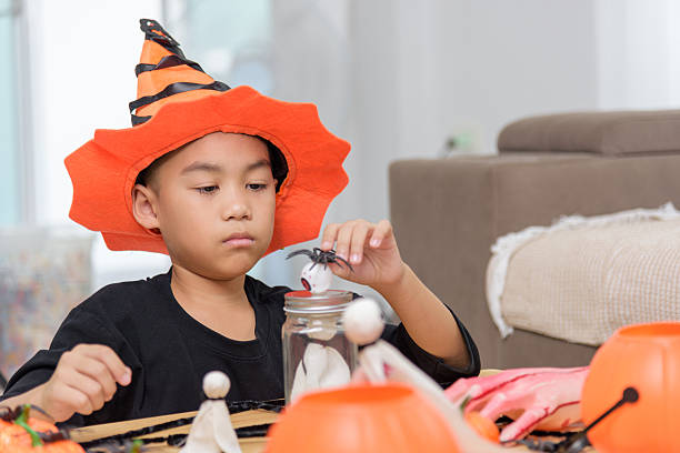 istock Boy in a costume hat is focused on arranging Halloween decorations like spiders and ghosts around a jar, getting ready for a spooky celebration at home 2233772683