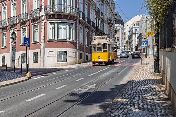 A yellow Lisbon tram travels down a street with traditional Portuguese buildings and a cobblestone sidewalk on a bright, sunny day. Lisbon, Portugal. 1 August 2025.