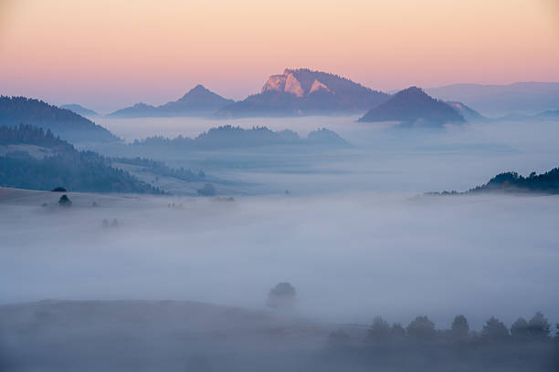Mountains are surrounded by thick fog at sunrise. A distant peak is illuminated by early light. The foreground is covered in mist.