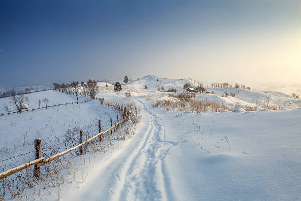 A snowy landscape features a path leading through the snow. A wooden fence runs alongside the pathway. Trees are scattered across the snow-covered hills.