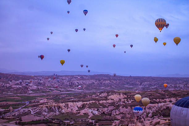 ​A stunning shot of hot air balloons soaring at sunrise over the surreal, rugged landscape of Cappadocia, Turkey. The morning light bathes the scene in a soft glow, highlighting the unique geological formations and the vibrant colors of the balloons.