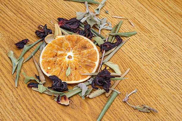 Dried a orange slice is centrally placed on wooden surface. Surrounded by an assortment of dried botanicals including dark red hibiscus flowers. Various ingredients for a herbal tea blend.