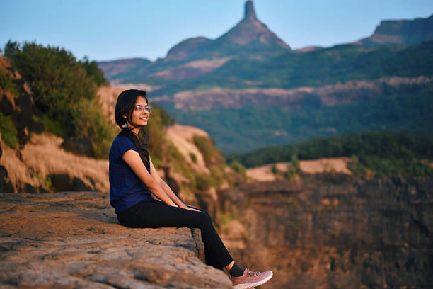 A young woman wearing glasses and a blue top sits on the edge of a rocky cliff, enjoying the view of the Sahyadri mountain range in Maharashtra, India. The scenic backdrop of rugged peaks and natural beauty, makes this an ideal concept for adventure, travel, exploration, and connecting with nature. Perfect for illustrating outdoor activities, freedom, mindfulness, or weekend getaways.