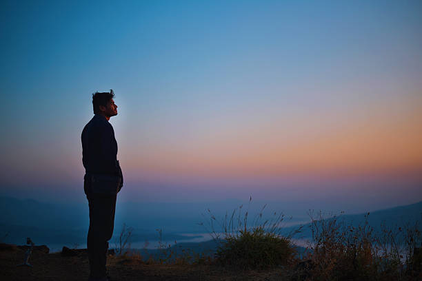 A man stands on the edge of a mountain cliff at dawn, gazing at the vibrant colors of the sunrise. The sky glows in shades of orange, pink, and blue, creating a peaceful and inspirational moment. Perfect for themes of solitude, adventure, mindfulness, travel, and natural beauty.