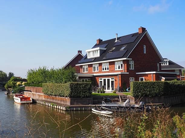 Leiden, Netherlands – September 10, 2023: A modern brick house with solar panels by a canal under a clear blue sky, featuring boats and lush greenery.