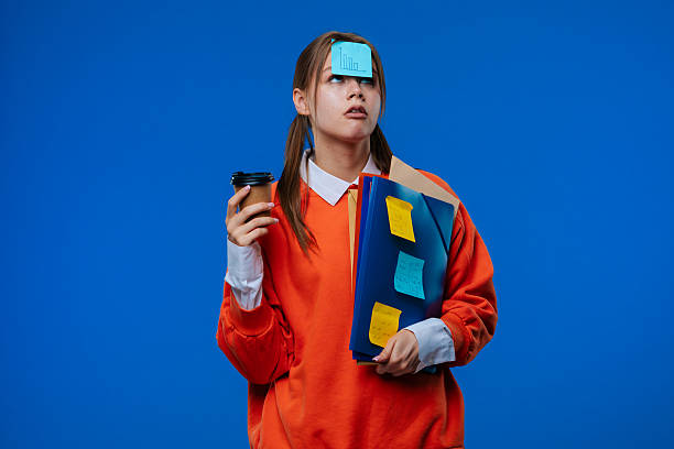 A young female student in an orange sweater poses with a sticker on her forehead, holding folders with notes and papers, to stand on a blue background. Education and student life.