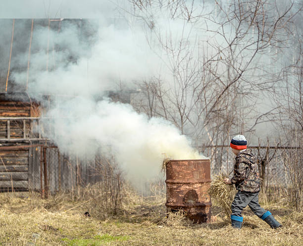 The cute boy is gathering dry grass and burning it in an old rusty barrel. Shooting in countryside at a homestead land in sunny spring day