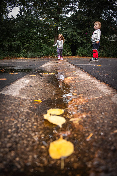Two sisters on a wet autumn road, one watching while the other runs near puddles after the rain. Seasonal childhood moment.