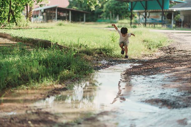 Asian boy leaps over a muddy puddle on a rural dirt path after rain. Wide composition with copy space, natural light, and clear action capture candid outdoor childhood energy and carefree fun.