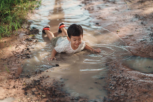 An Asian boy lies and slides through a muddy puddle on a dirt path after rain, smiling in warm natural light. Wide, candid framing with copy space conveys messy play, sensory learning, and joy.