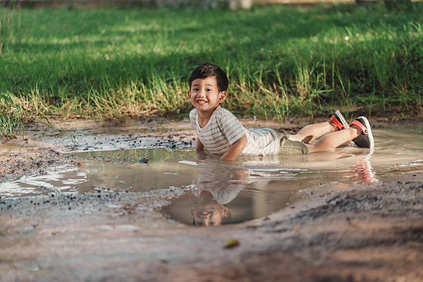 Smiling Asian boy lies in a muddy puddle on a rural dirt path after rain. Natural light, reflection, and wide copy space capture candid outdoor childhood, messy sensory play, and carefree joy.