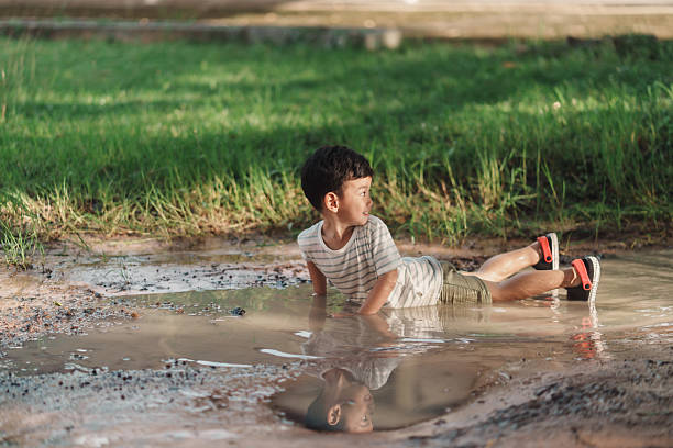 Asian boy lies in a muddy puddle on a rural dirt path after rain. Natural light, and wide copy space highlight candid outdoor childhood, messy sensory play, and carefree curiosity in nature