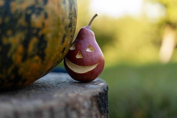 Red pear with halloween face carved on its skin, near a pumpkin, resting on a tree stump in a garden
