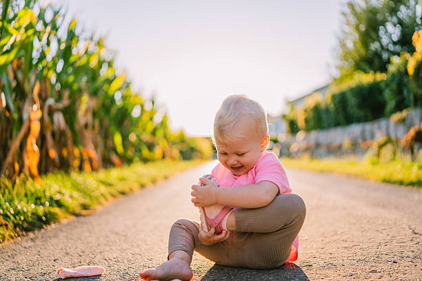Small child sitting cross-legged on a paved road holding a shoe, smiling, with greenery and cornfield in the background in Germany.