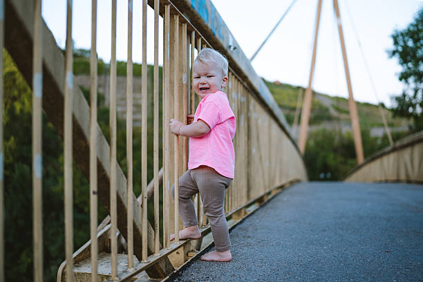 Young child barefoot, standing on pedestrian bridge holding railing and looking back. Outdoor candid lifestyle photo in Germany.