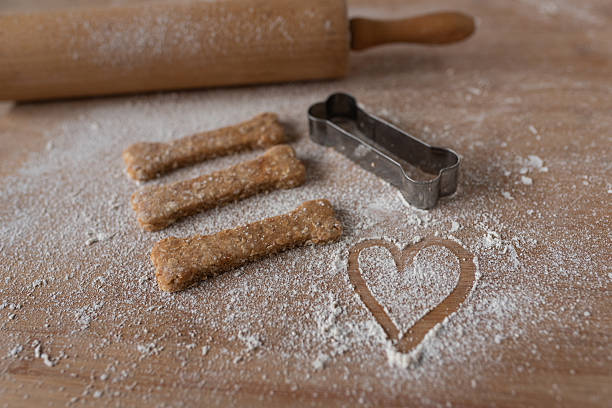 Homemade dog biscuits on a floured wooden surface with a rolling pin, bone-shaped cookie cutter and a heart drawn in flour.