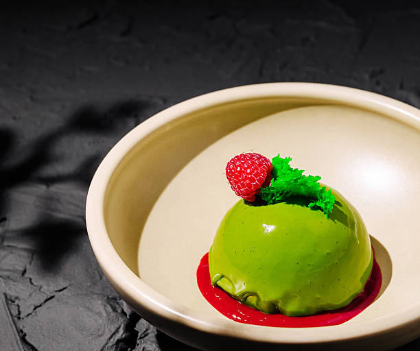 A vibrant green dessert sits in a simple bowl, topped with a fresh raspberry and mint leaf. The dessert rests on a smooth, dark surface, enhancing its visual appeal.
