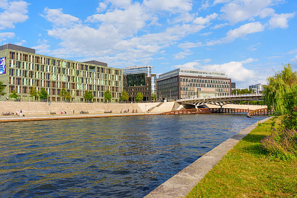 Berlin, Germany - August 20, 2025: Contemporary buildings reflect in the Spree River while people relax along the waterfront pathway under a blue sky with scattered clouds.