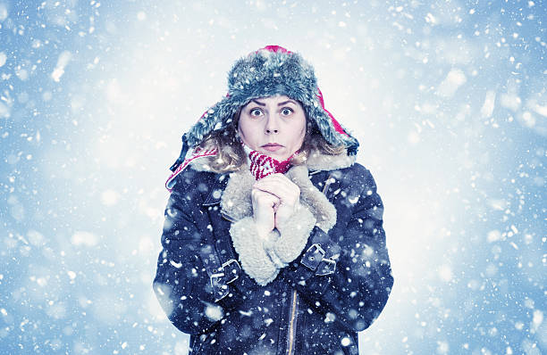 Frozen woman in winter coat, fur hat and scarf standing outdoors in snowfall. She looks cold, shivering, holding hands close to face. Concept of freezing weather, snowstorm, low temperature. stock photo