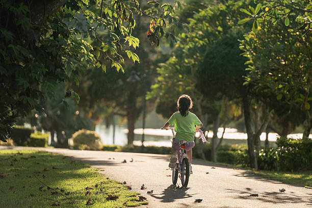 Child Riding Bicycle Through Lush Green Park Pathway Under Soft Sunlight Surrounded by Beautiful Trees and Nature in the Early Evening
