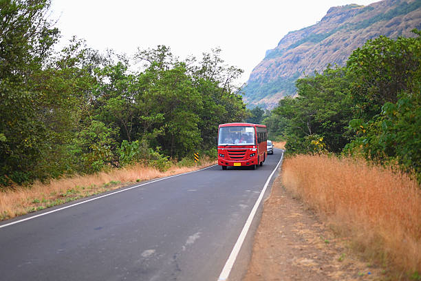 A red bus travels along a winding mountain road surrounded by lush green trees and dry golden grass, with rocky hills in the background. Capturing a moment of travel through the countryside. Perfect for themes of transportation, rural journeys, or scenic road trips.