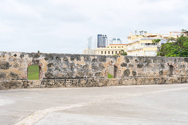 Weathered stone wall with archways stands before a modern city skyline under a cloudy sky