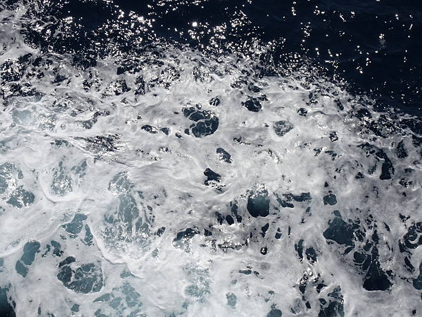 Dynamic close-up of ocean waves with white sea foam and sparkling sunlight reflections on deep blue water. The abstract pattern of bubbles and liquid movement captures the energy and natural texture of the sea, perfect for themes of freshness, marine life, water background, and nature's powe