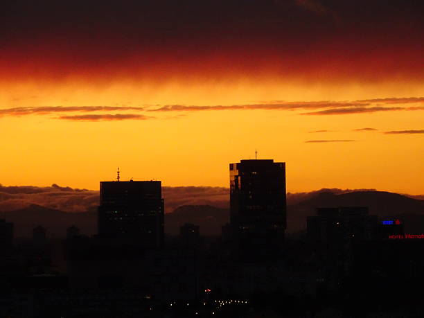 Bold silhouette of modern urban high-rise buildings set against a vibrant, gradient sunset sky in tones of orange, yellow, and deep red. The striking colors and layered clouds create a powerful, atmospheric backdrop conveying urban life, evening stillness, and cityscape beauty. Ideal for concepts like architecture, city living, sunset, and dramatic urban perspectives