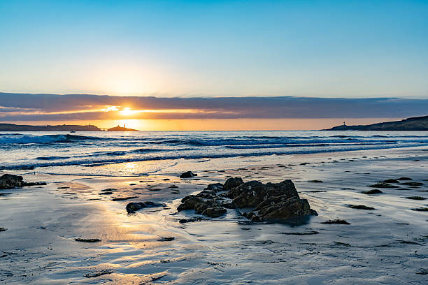 Asturias province. Spain. Golden sunrise reflecting on wet sand and gentle ocean waves, with two distant lighthouses adding a touch of serenity to the tranquil coastal scene