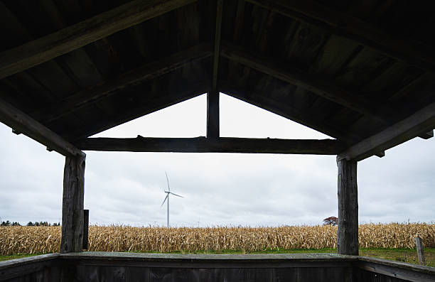 A serene rural scene featuring a wooden structure framing a view of a field with a wind turbine, all under a cloudy sky providing a tranquil and sustainable ambiance.