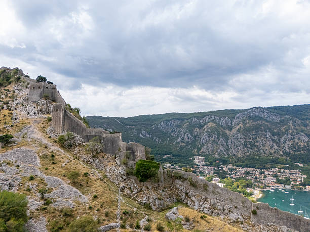 Historic stone fortress perched on rocky hillside above a coastal town with boats in the bay. Scenic mountain landscape with cloudy sky in the background. Old architecture and nature.