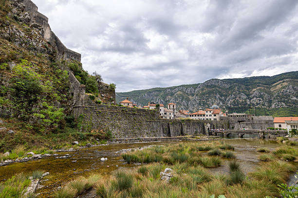 Ancient stone fortress walls with a stone bridge over a shallow river surrounded by grass and hills. Old European town architecture with mountains in the background. Scenic historic landscape view.