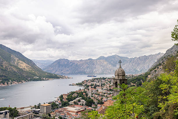 Stone church tower with cross beside narrow stone path on hillside. Scenic coastal town with red rooftops and fjord surrounded by mountains. Mountain landscape with cloudy sky.