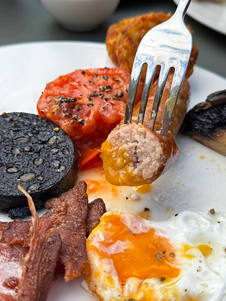 Close-up image of full English fried breakfast on white plate, metal  fork stuck in sausage, bacon rasher, grilled tomato half, slice of black pudding, sunny-side-up fried egg, hash brown and portobello mushroom, elevated view, focus on foreground