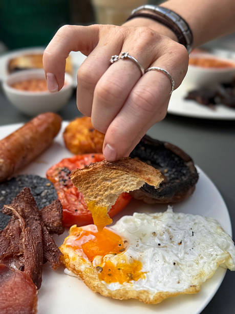 Close-up image of full English fried breakfast on white plate, unrecognisable person dipping white toast in yolk of sunny-side-up fried egg, sausage, bacon rasher, grilled tomato half, slice of black pudding, hash brown and portobello mushroom