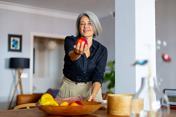 istock Senior woman offering apple while preparing fruit bowl in dining room 2233918319