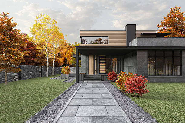 Contemporary two-story residential home with sleek architectural design. The structure features a harmonious blend of natural wood paneling, dark stone or concrete textures, and expansive glass windows that invite natural light into the interior. The house is characterized by its flat roof, clean geometric lines, and minimalist aesthetic. A black overhang supported by a column marks the covered entrance, while large stone tiles form a pathway leading to the front door. The surrounding landscaping includes manicured green grass, ornamental shrubs, and trees adorned with vibrant autumn foliage, adding warmth and seasonal charm to the scene. A raised platform with steps and a side garage complete the layout, enhancing both functionality and visual appeal.\n\nThis image is ideal for use in architectural portfolios, real estate listings, construction and design websites, home improvement blogs, or promotional materials for modern housing developments. It can also serve as inspiration for exterior design, landscaping concepts, or eco-friendly building projects. The combination of modern architecture and seasonal ambiance makes it perfect for showcasing upscale living, suburban tranquility, or innovative residential design.