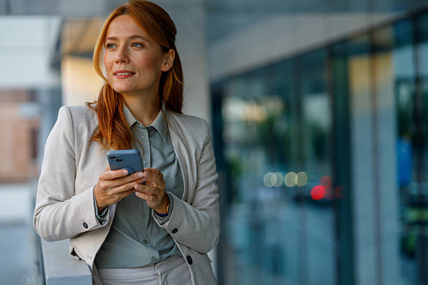 istock Businesswoman is holding smartphone and looking away while leaning on railing in front of office building 2233920182