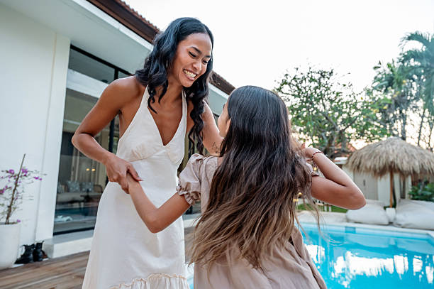 A joyful moment of a mother and daughter spending quality time outdoors near a pool, enjoying laughter and bonding amidst a serene tropical backdrop during a sunny day.