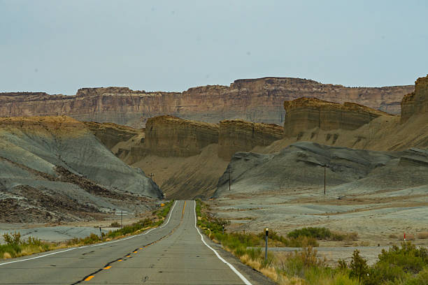 An expansive view of an empty, two-lane asphalt highway stretching into the distance, with double yellow lines marking its path through a dramatic and rugged desert landscape. On either side of the road, unique badlands formations rise, showcasing distinct layers of grey, tan, and brown sedimentary rock, hinting at ancient geological processes and erosion. In the background, a towering mesa or plateau forms a majestic and textured backdrop under a soft, overcast sky, contributing to a sense of vastness and solitude. The composition utilizes the road as a strong leading line, drawing the viewer's eye deep into the scene, emphasizing the journey ahead. This powerful image evokes feelings of adventure, freedom, and the allure of the open road, making it ideal for travel narratives, environmental studies, themes of exploration, or concepts of finding a path forward across remote and challenging landscapes, characteristic of the American Southwest.