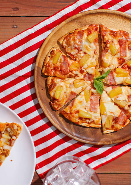 Top view of fast food delicious pineapple and ham hawaiian pizza with mozzarella cheese on red and white striped placemat on wooden table, homemade cooking ingredients on dark background advertisement