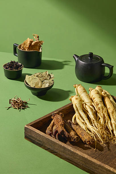 Vertical photo with ginseng roots standing on a tray beside a tea set and herbs on green background. A traditional and natural look for wellness themes.