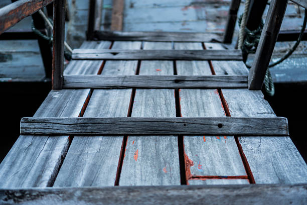 A low-angle shot of a worn, weathered wooden gangplank with visible streaks of faded blue and red paint. The rustic texture of the planks creates a sense of history and nautical adventure, leading the viewer's eye toward a boat at a pier, just out of view.