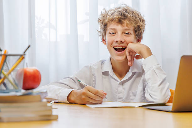 istock Schoolboy studying at home with laptop, notebook, and learning materials, smiling happily 2233971394