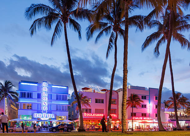 Miami Beach. USA. 04.14.2025. Evening view of Ocean Drive with neon-lit hotels, palm trees, vintage cars and walking tourists in Miami Beach, Florida.