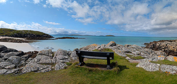 This coastal image from Eriskay in the Outer Hebrides, Scotland, features a bench overlooking turquoise waters, a sandy beach, and distant islands under a partly cloudy summer sky. Captured in August, the scene evokes tranquility, reflection, and natural beauty. Ideal for themes in travel, mindfulness, and environmental awareness.